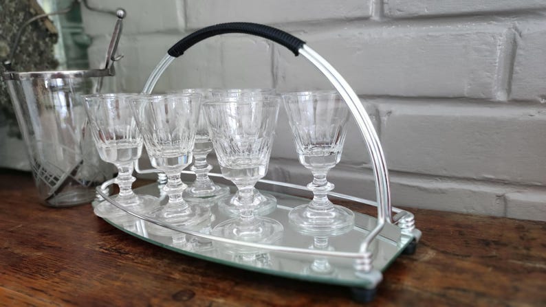 May include: A silver serving tray with six clear, stemmed glasses. The tray has a mirrored surface and a curved handle with a black grip. An ice bucket is visible in the background. The glasses are approximately 4 inches tall.