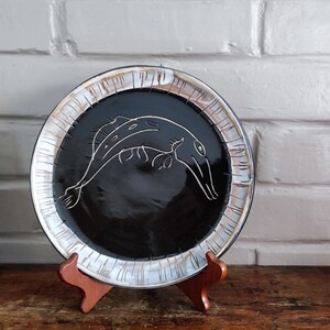 Peut inclure: Une assiette décorative avec un centre noir et un contour blanc. Un dessin au trait blanc d'un poisson est au centre. L'assiette est présentée sur un support en bois. L'assiette est un objet décoratif.
