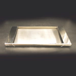 Op de afbeelding: Een rechthoekig zilverkleurig metalen dienblad met een gepolijste, reflecterende afwerking. Het dienblad heeft een plat centraal oppervlak dat wordt begrensd door twee brede, gladde en gebogen zijpanelen aan de linker- en rechterkant.