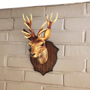 Op de afbeelding: Een aan de muur gemonteerde hertenkopsculptuur met een beige en bruine afwerking en gedetailleerd gewei. De hertenkop is bevestigd op een donkerbruine, schildvormige plaat met houtnerftextuur voor decoratie aan de muur.