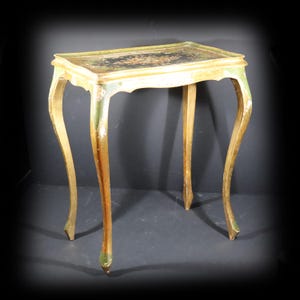 Peut inclure: Une table d'appoint rectangulaire dotée de quatre pieds galbés, présentant une finition peinte en vert et doré vieilli. Le plateau comporte un panneau central sombre orné d'un motif floral, encadré par un bord doré surélevé.