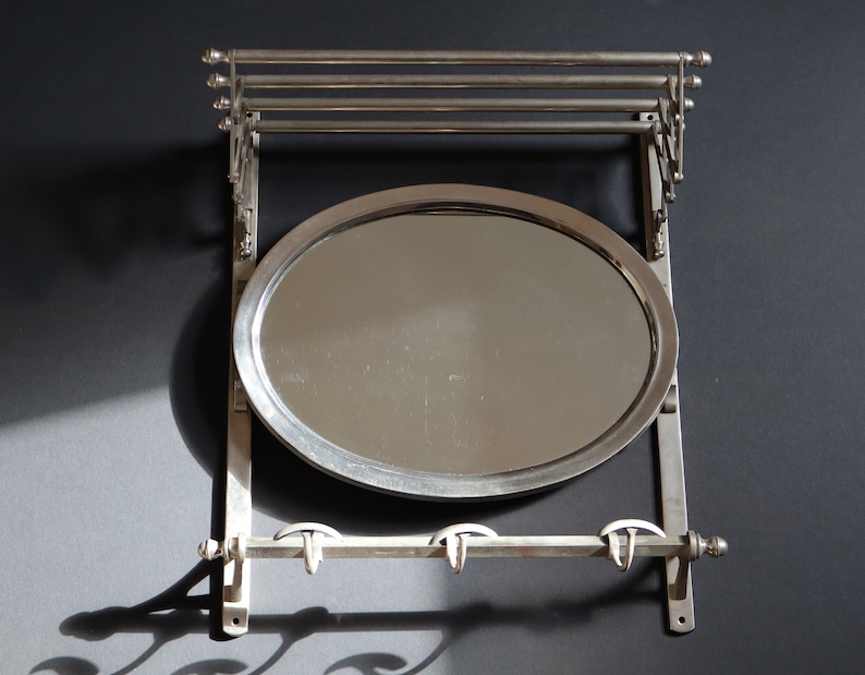 May include: A silver wall-mounted coat rack with a round mirror and three hooks. The rack has a decorative design with scrolls and curves.