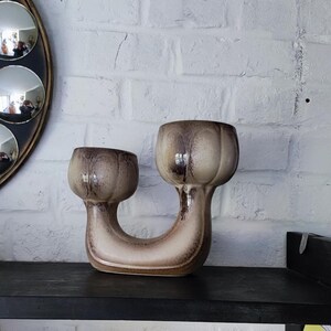 May include: A ceramic, U-shaped vase with two bulbous openings. The vase is a mix of beige and brown tones, with darker brown accents. It sits on a black shelf against a white brick wall. The vase is a decorative item.