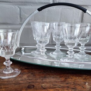 May include: A set of six clear crystal cordial glasses with a decorative etched design, displayed on a mirrored tray with a silver metal handle. The glasses are on a wooden surface.