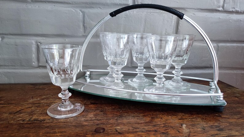 May include: A set of six clear crystal cordial glasses with a decorative etched design, displayed on a mirrored tray with a silver metal handle. The glasses are on a wooden surface.