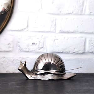 May include: A metallic silver snail-shaped serving dish with a rounded, segmented shell that functions as a lid. The snail has a smooth, reflective finish and includes a small, thin metal spoon resting against the side of the shell body.
