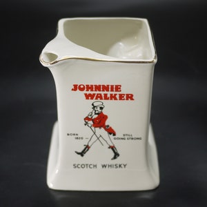May include: White ceramic pitcher with a gold rim, featuring the "Johnnie Walker" logo in red. The logo includes a drawing of a man in a red coat and the words "Scotch Whisky". The pitcher has a unique, angular design.