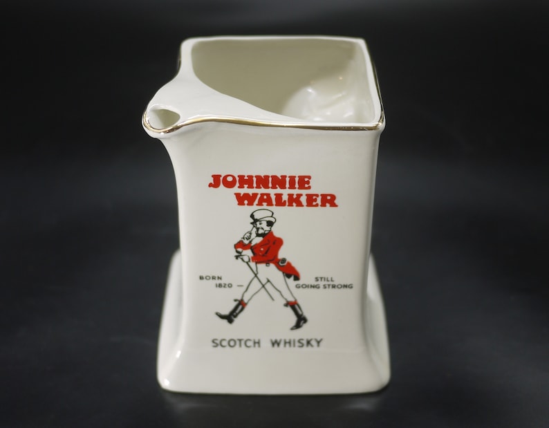 May include: White ceramic pitcher with a gold rim, featuring the "Johnnie Walker" logo in red. The logo includes a drawing of a man in a red coat and the words "Scotch Whisky". The pitcher has a unique, angular design.