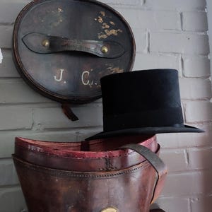 May include: A black top hat resting on a brown leather hat box. The cylindrical hat box features a brass lock on the front, a leather handle on the lid, and the initials J. C. painted in white on the lid. The interior of the box is lined in red.
