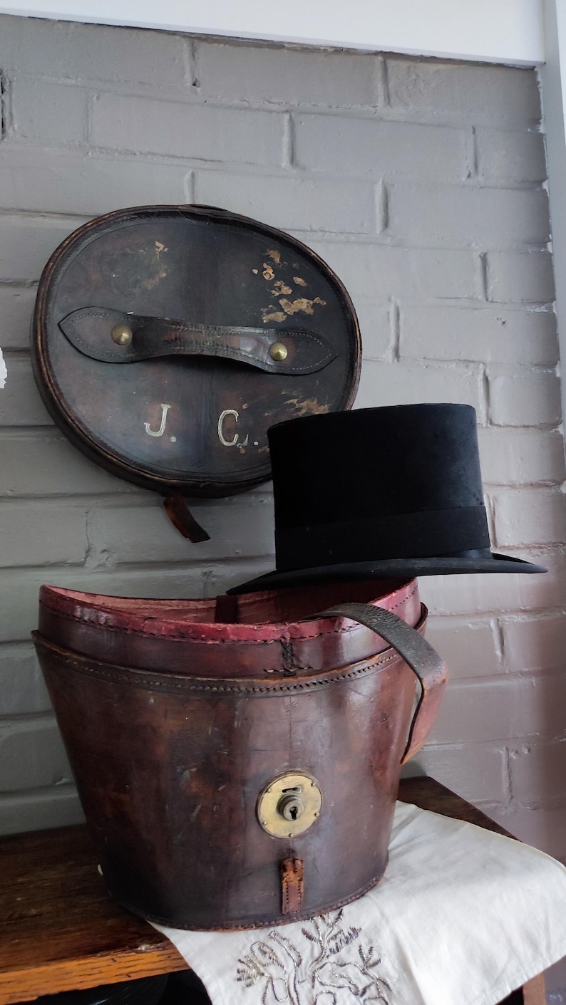 May include: A black top hat resting on a brown leather hat box. The cylindrical hat box features a brass lock on the front, a leather handle on the lid, and the initials J. C. painted in white on the lid. The interior of the box is lined in red.
