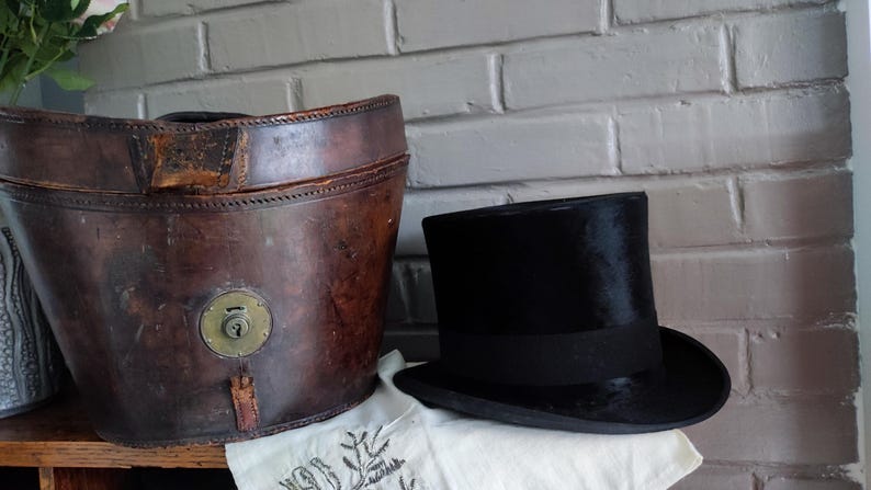 May include: A black top hat with a matching black ribbon band positioned next to a large, oval-shaped brown leather hat box. The leather container features a prominent circular brass lock mechanism on the front and visible stitched seams.