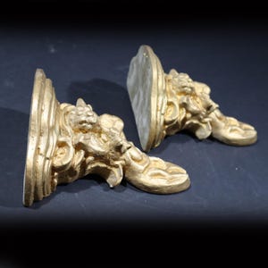 May include: A pair of gold-colored decorative wall brackets with ornate designs. The brackets are made of a lightweight material and have a distressed finish.