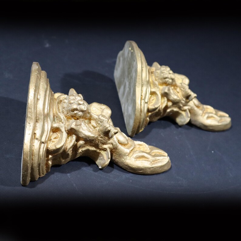 May include: A pair of gold-colored decorative wall brackets with ornate designs. The brackets are made of a lightweight material and have a distressed finish.