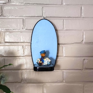 Peut inclure: Figurine vintage d'un policier à moto, exposée sur une étagère noire sur fond bleu clair. La figurine porte un chapeau et un uniforme bleus. L'étagère est fixée sur un mur de briques blanches.