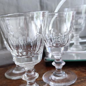 May include: Clear, stemmed glassware with etched designs. The glasses have a rounded bowl, a decorative stem, and a circular base. The glassware is likely for serving beverages. The glasses are on a wooden surface.