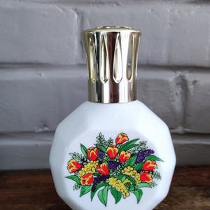 Peut inclure: Une lampe à parfum en verre blanc à facettes ornée d'un motif floral coloré représentant des tulipes rouges, du mimosa jaune et des fleurs violettes. La lampe est surmontée d'un capuchon brûleur doré en forme de couronne ajourée.