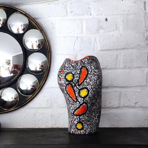 Peut inclure: Un vase en céramique mouchetée avec un motif abstrait de formes orange et jaunes soulignées de noir. Le vase a une surface texturée et un bord orange. Un miroir rond avec plusieurs miroirs convexes est en arrière-plan.