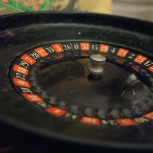 May include: A black roulette wheel featuring a central silver knob and a circular track with alternating red and black numbered segments. The numbers are printed in white, and the wheel includes a zero, with a smooth, dark, circular outer rim.