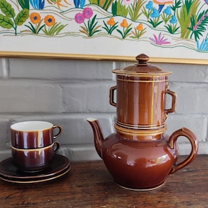 Peut inclure: Un service à café vintage en céramique marron comprenant une cafetière, deux tasses et des soucoupes. La cafetière a un design unique avec un dessus cylindrique et une base arrondie. Les tasses et les soucoupes sont de couleur marron assortie avec un intérieur blanc.
