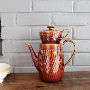 Op de afbeelding: Een bruine keramische koffiepot met een bijpassende creamer. De koffiepot heeft een hoge, cilindrische vorm met een gebogen handvat en tuit. De creamer staat bovenop de pot. Beide stukken hebben een geribbeld, gedraaid ontwerp.