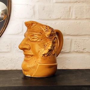 Peut inclure: Une cruche en céramique émaillée jaune moutarde, représentant le profil d'un visage humain avec un nez proéminent, des cheveux texturés et des favoris. L'objet possède une anse incurvée sur le côté droit et un col moulé à la base.