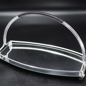 May include: A silver-toned serving tray with a curved handle featuring a black, textured grip. The tray has a mirrored surface and rests on small, rounded feet. The design is sleek and modern, suitable for serving food or displaying items.