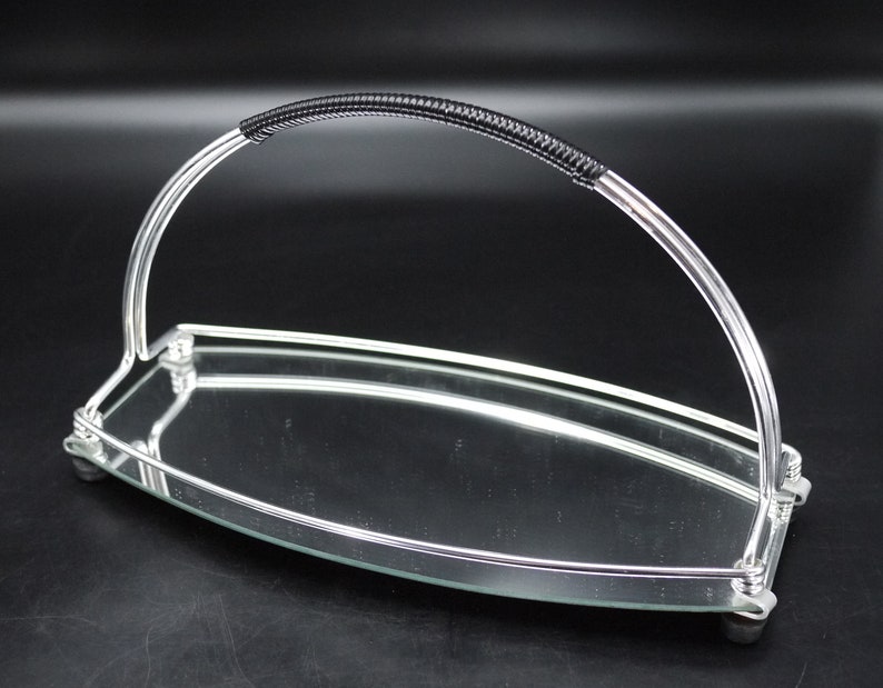 May include: A silver-toned serving tray with a curved handle featuring a black, textured grip. The tray has a mirrored surface and rests on small, rounded feet. The design is sleek and modern, suitable for serving food or displaying items.