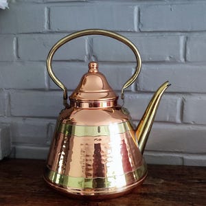 May include: A copper tea kettle with a hammered finish and brass accents. The kettle has a curved spout and a brass handle. The kettle is sitting on a wooden surface. The background is a white brick wall.