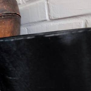 May include: A black top hat with a tall, cylindrical crown and a flat top, positioned next to a large, weathered brown leather container with visible stitching along its curved surface and a textured, aged appearance.