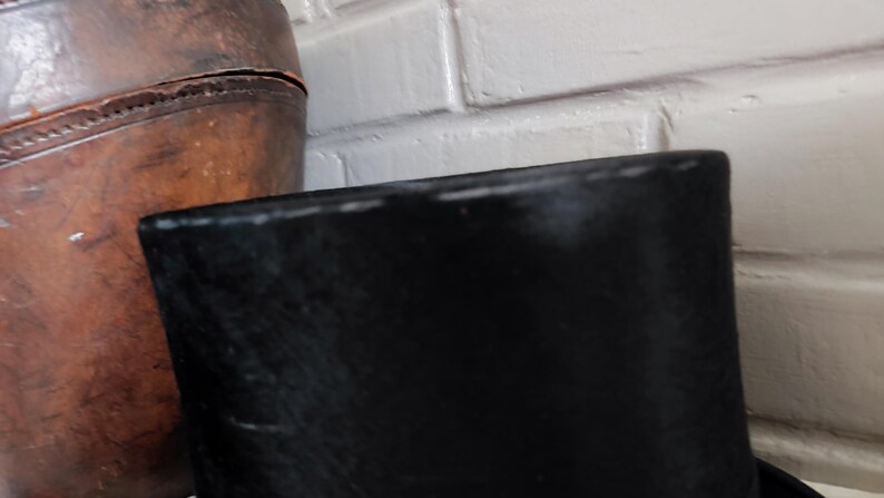 May include: A black top hat with a tall, cylindrical crown and a flat top, positioned next to a large, weathered brown leather container with visible stitching along its curved surface and a textured, aged appearance.
