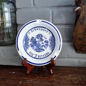 May include: A round white ceramic ashtray featuring a blue willow pattern illustration in the center. The text Schweppes and THE 3 BITTERS is printed in blue. The ashtray has a blue rim and three notches for resting cigarettes along the edge.