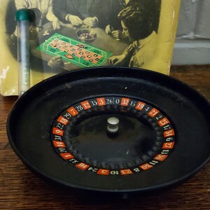 May include: A black roulette wheel with a central silver knob and a numbered ring featuring red, black, and green segments. Behind the wheel sits a yellow game box cover depicting people playing a board game, alongside a clear plastic tube.