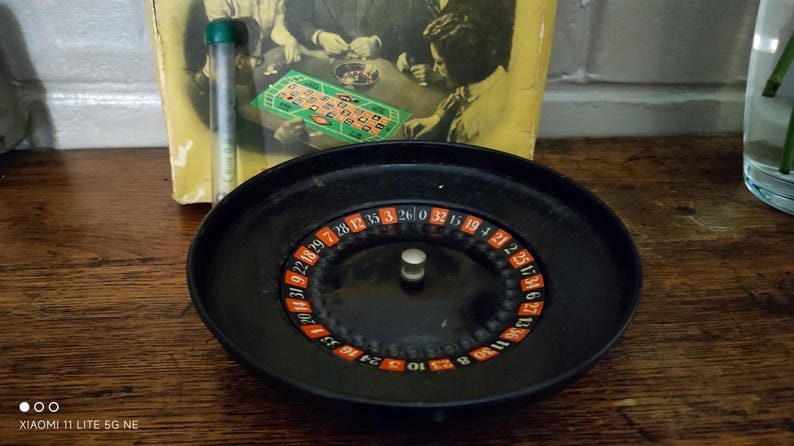 May include: A black roulette wheel with a central silver knob and a numbered ring featuring red, black, and green segments. Behind the wheel sits a yellow game box cover depicting people playing a board game, alongside a clear plastic tube.