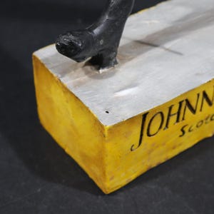 May include: A sculpture featuring a black figure on a silver surface, set on a yellow block. The words "JOHNN" and "Scotc" are written in black script on the yellow block.