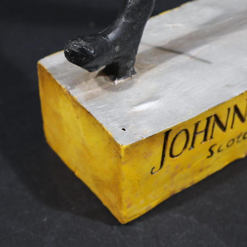 May include: A sculpture featuring a black figure on a silver surface, set on a yellow block. The words "JOHNN" and "Scotc" are written in black script on the yellow block.