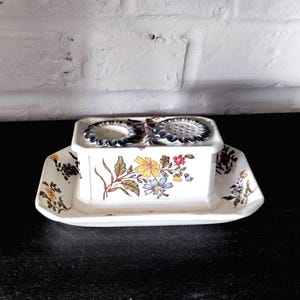 May include: A white ceramic butter dish with a floral design featuring yellow, blue, and red flowers. The dish has a rectangular base and a lid with two circular openings. The edges are scalloped and the dish sits on a black surface.