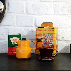 May include: A vintage Melitta Kinder-Filter-Set featuring a bright orange ceramic coffee pot with a dark brown lid, a dark brown ceramic coffee filter cone, and original packaging boxes with text including Melitta and Kinder-Filter-Set.