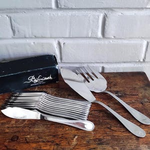 May include: A set of silver-toned fish cutlery including a serving knife and fork, alongside a stack of smaller fish knives. The black box is labeled with the text Béfinoix Orfèvrerie de Table. The utensils feature a polished finish and simple handles.