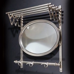 May include: A silver wall-mounted coat rack with a mirror and a fold-down towel rack. The rack has three hooks and a round mirror.