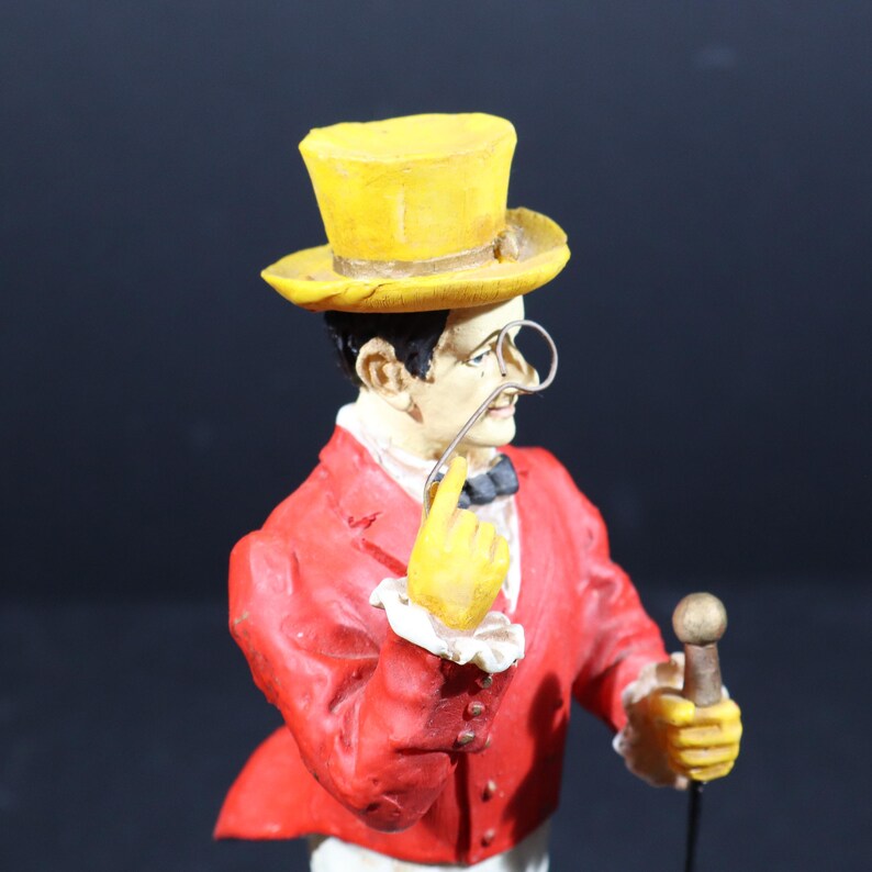 May include: A figurine of a man in a red jacket, yellow top hat, and yellow gloves, holding a cane. The man has a monocle and a bow tie. The figurine is a decorative item.