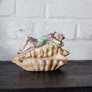 Puede incluir: Figura de porcelana de una mujer reclinada sobre una gran concha. La mujer lleva un traje de baño rosa, verde y blanco y tiene el pelo rubio. La concha es beige y tiene una superficie texturizada. La figura es un objeto decorativo.