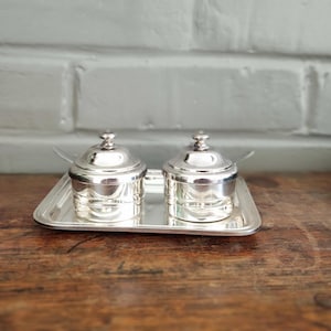 Peut inclure: Un ensemble de deux petits pots à condiments argentés polis avec couvercles assortis et petites cuillères, disposés sur un plateau rectangulaire argenté. Chaque pot présente un corps cylindrique lisse avec un couvercle bombé et un bouton.