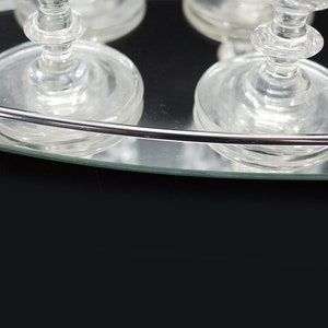 May include: Clear glass stemware on a mirrored tray with a silver rim. The glasses have a classic design with a round base and a detailed stem. The tray reflects the glasses and the dark background.