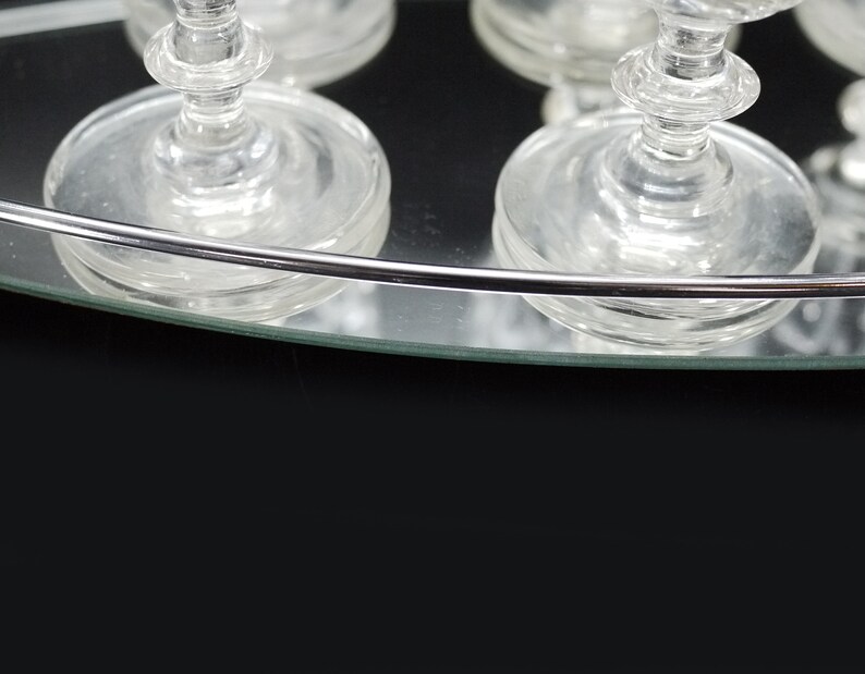 May include: Clear glass stemware on a mirrored tray with a silver rim. The glasses have a classic design with a round base and a detailed stem. The tray reflects the glasses and the dark background.