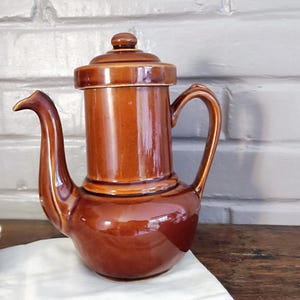 Peut inclure: Une cafetière en céramique marron avec un couvercle, une poignée et un long bec verseur. Le pot est posé sur un tissu blanc à côté d'une tasse à thé blanche avec un bord doré et un motif floral. L'arrière-plan est un mur de briques blanches.