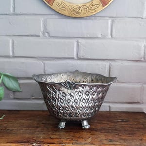 Könnte beinhalten: Eine silberfarbene Metallschale mit einer erhabenen Rautenmuster-Textur auf der Außenseite, einem gewellten Rand mit dekorativen Details und drei kleinen Füßen an der Basis. Die Schale hat eine weite Öffnung und eine polierte, reflektierende Oberfläche.