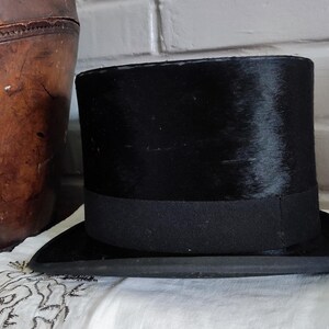 May include: A black top hat with a flat crown and a wide, curved brim. The hat features a smooth, dark finish and a matching black grosgrain ribbon band encircling the base of the crown, providing a structured and uniform appearance throughout.