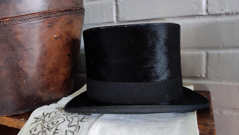 May include: A black top hat with a flat crown and a wide, curved brim. The hat features a smooth, dark finish and a matching black grosgrain ribbon band encircling the base of the crown, providing a structured and uniform appearance throughout.