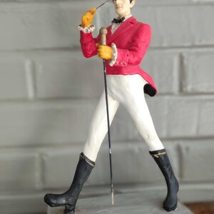 May include: A Johnnie Walker Striding Man figurine. The figure wears a red jacket, white trousers, and black boots, and holds a cane. The base is a yellow rectangle with the words "Johnnie Walker Scotch Whisky".