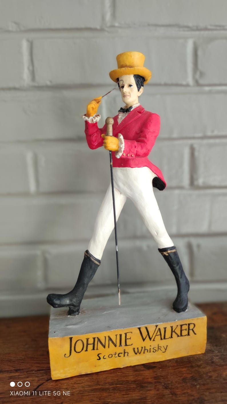 May include: A Johnnie Walker Striding Man figurine. The figure wears a red jacket, white trousers, and black boots, and holds a cane. The base is a yellow rectangle with the words "Johnnie Walker Scotch Whisky".